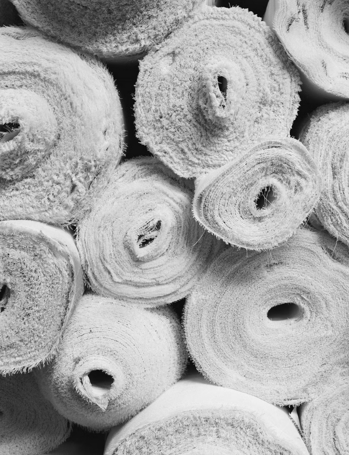 Black and white photo of multiple rolled fabric or carpet materials stacked together, showing circular spiral patterns and frayed edges.