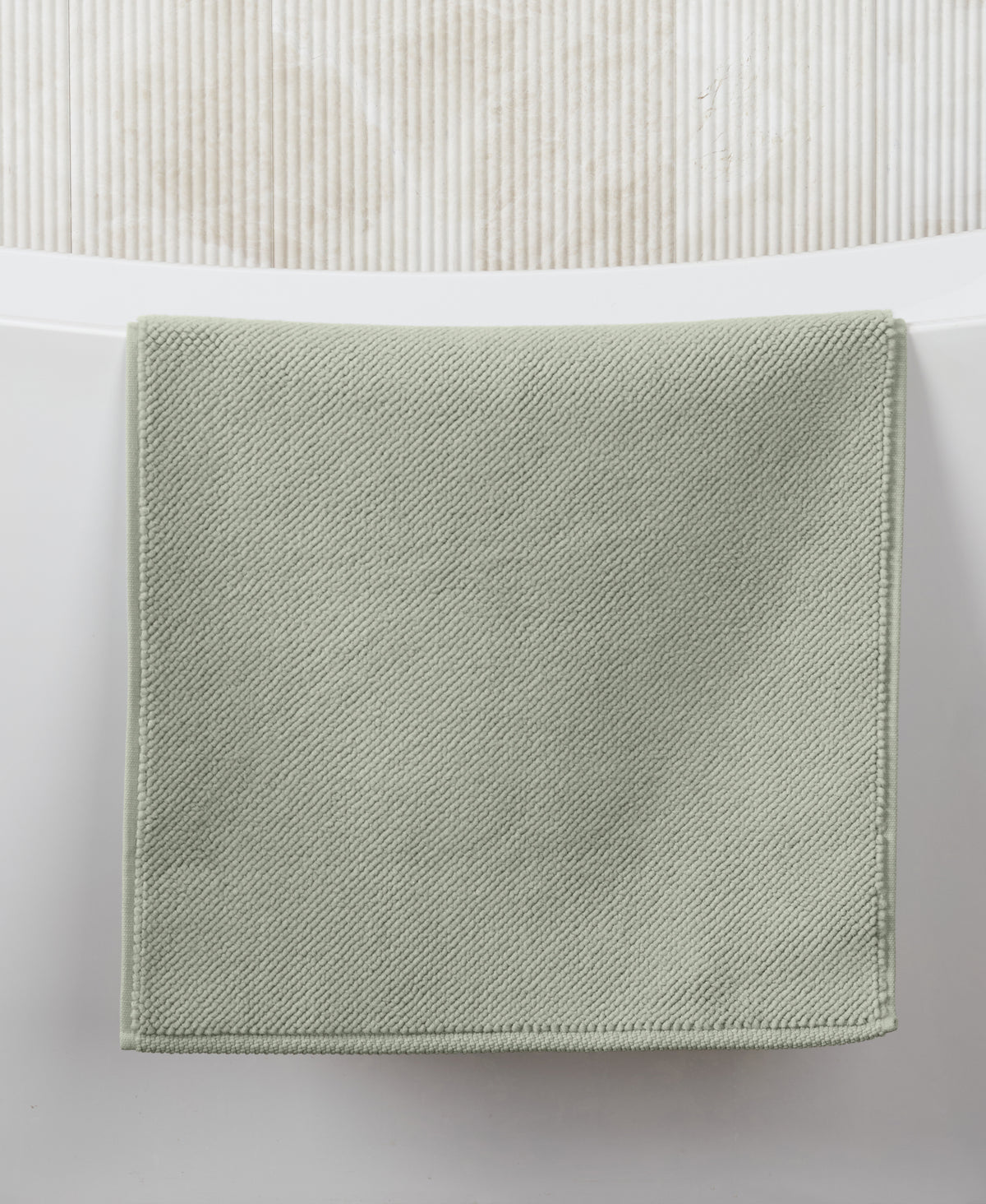 Sage green square bath mat with textured diagonal weave pattern on white tiled bathroom floor