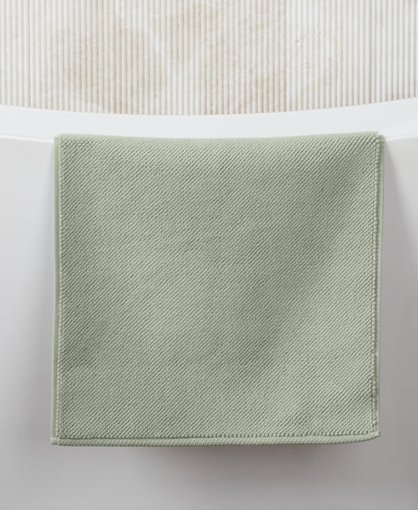 Sage green square bath mat with textured diagonal weave pattern on white tiled bathroom floor