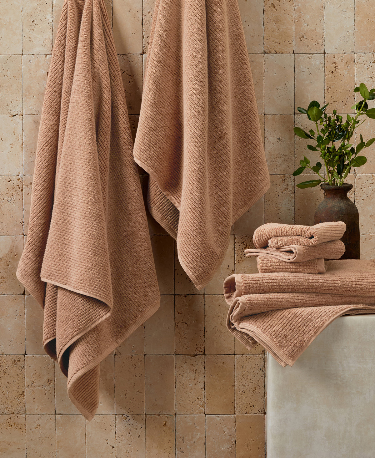 Adobe-colored ribbed towels hanging on travertine wall with folded towels stacked on white surface and plant in ceramic vase