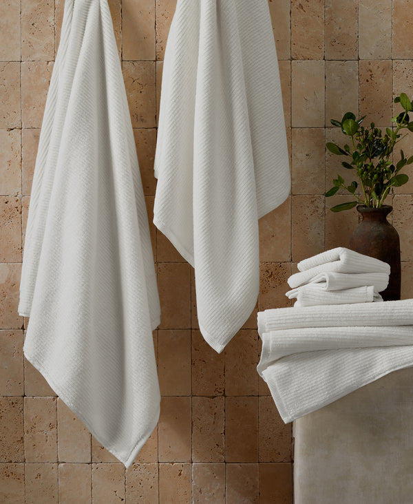 White ribbed towels hanging on hooks and stacked on shelf against beige travertine tile wall with decorative vase
