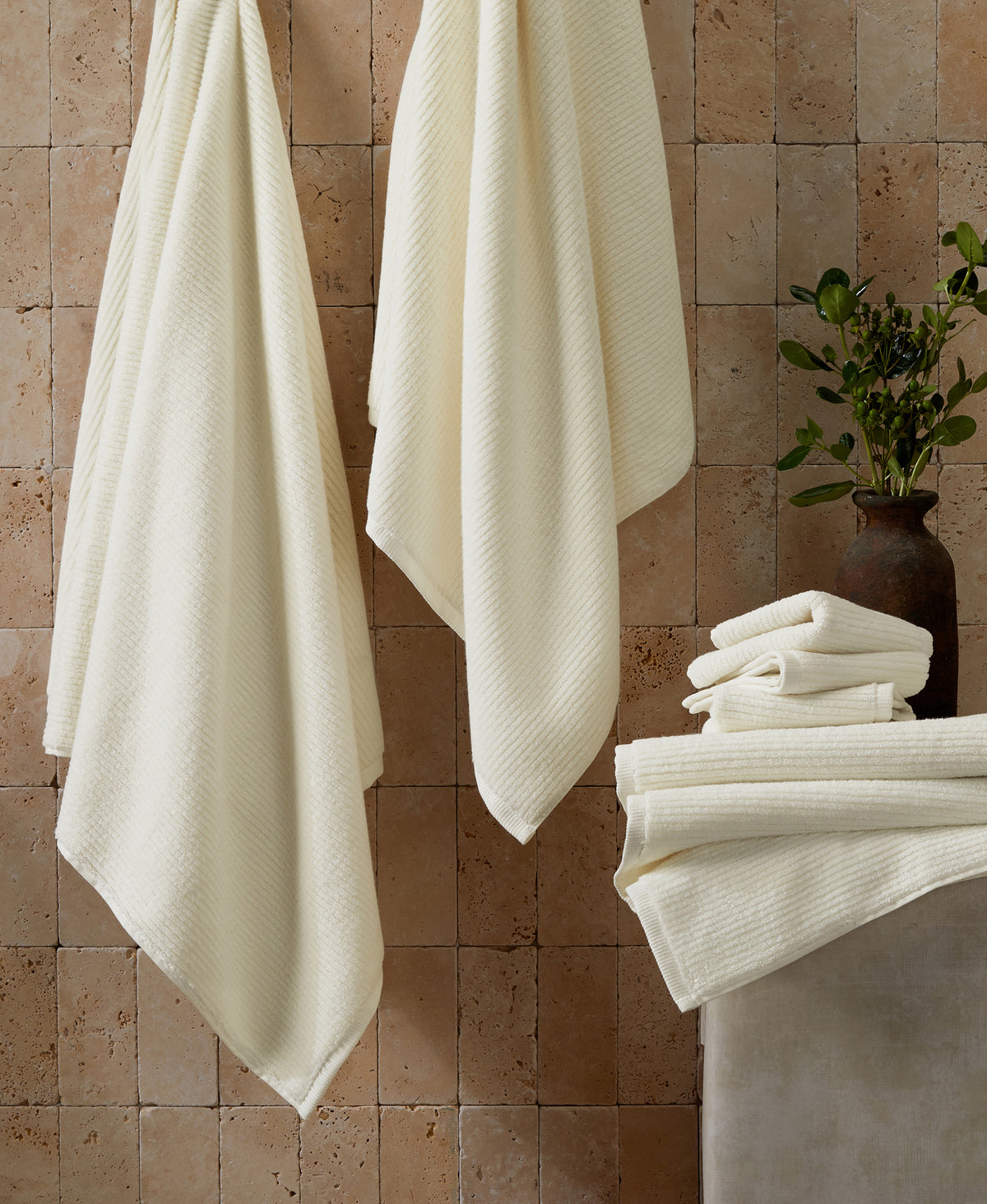 Cream-colored ribbed towels hanging on hooks and stacked on ledge against travertine tile wall with potted plant