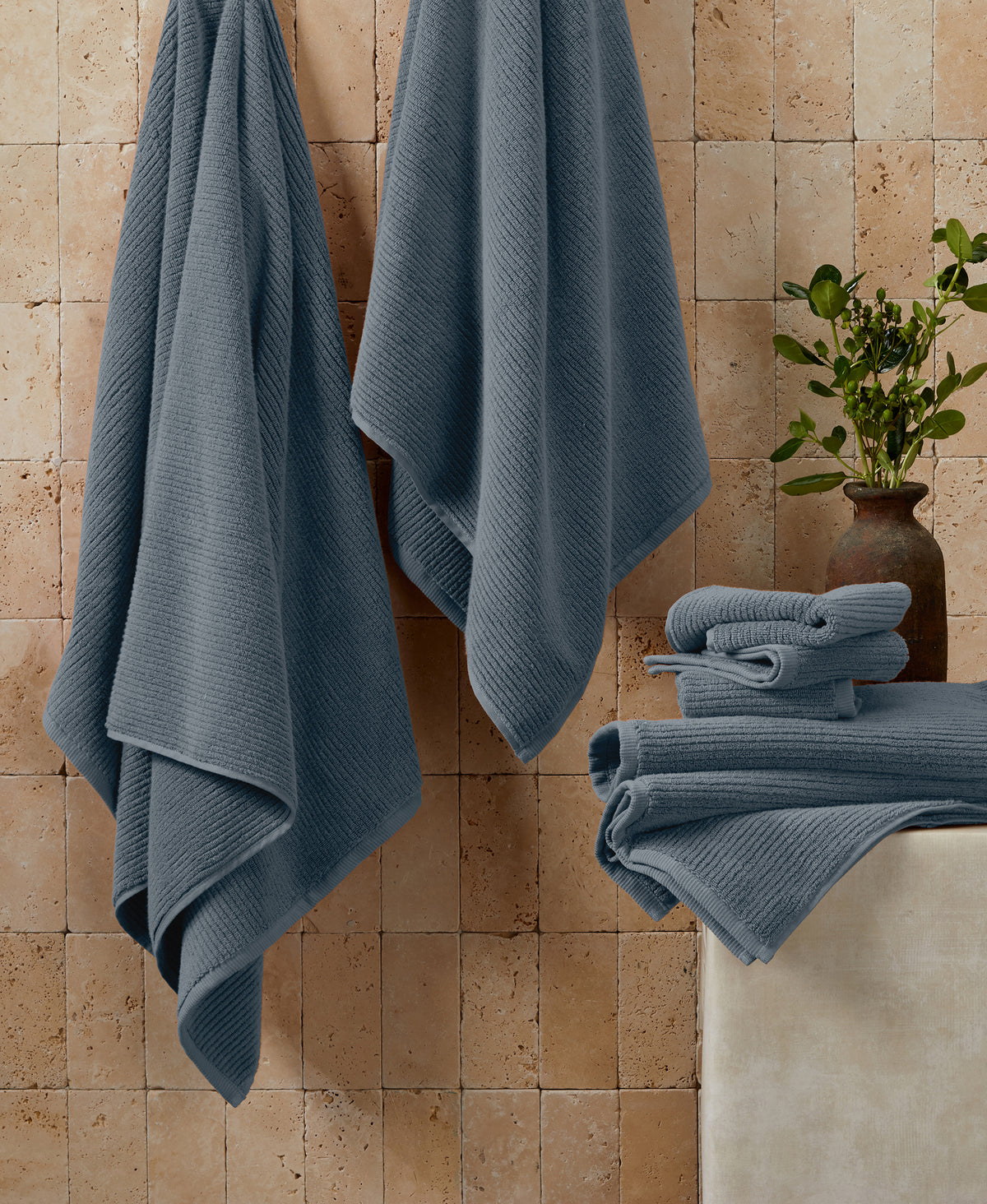 Blue ribbed towels hanging on wall with folded towels on bathtub edge, travertine tile background