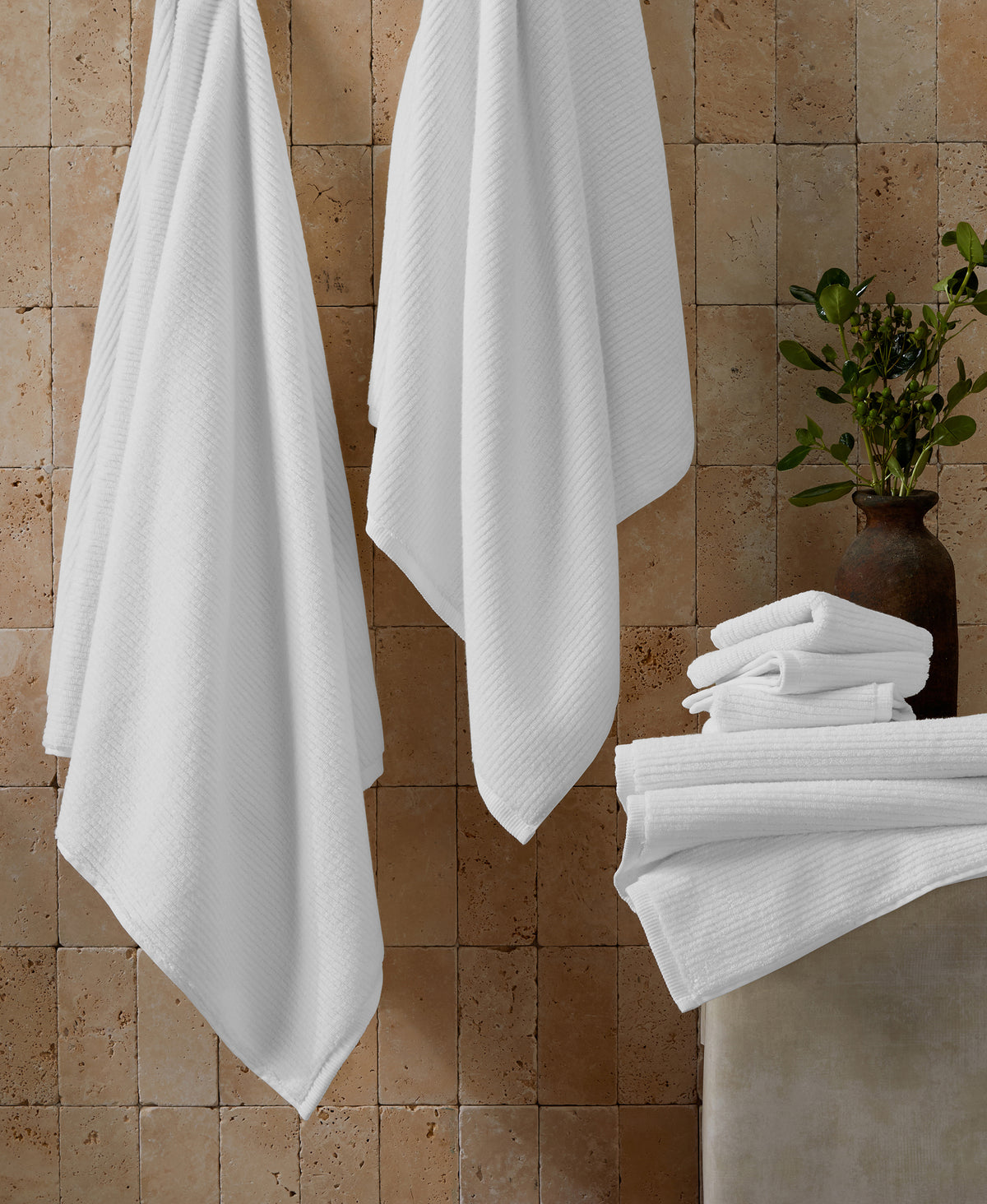 White ribbed towels hanging on hooks and folded in stack against textured stone wall with plant in vase