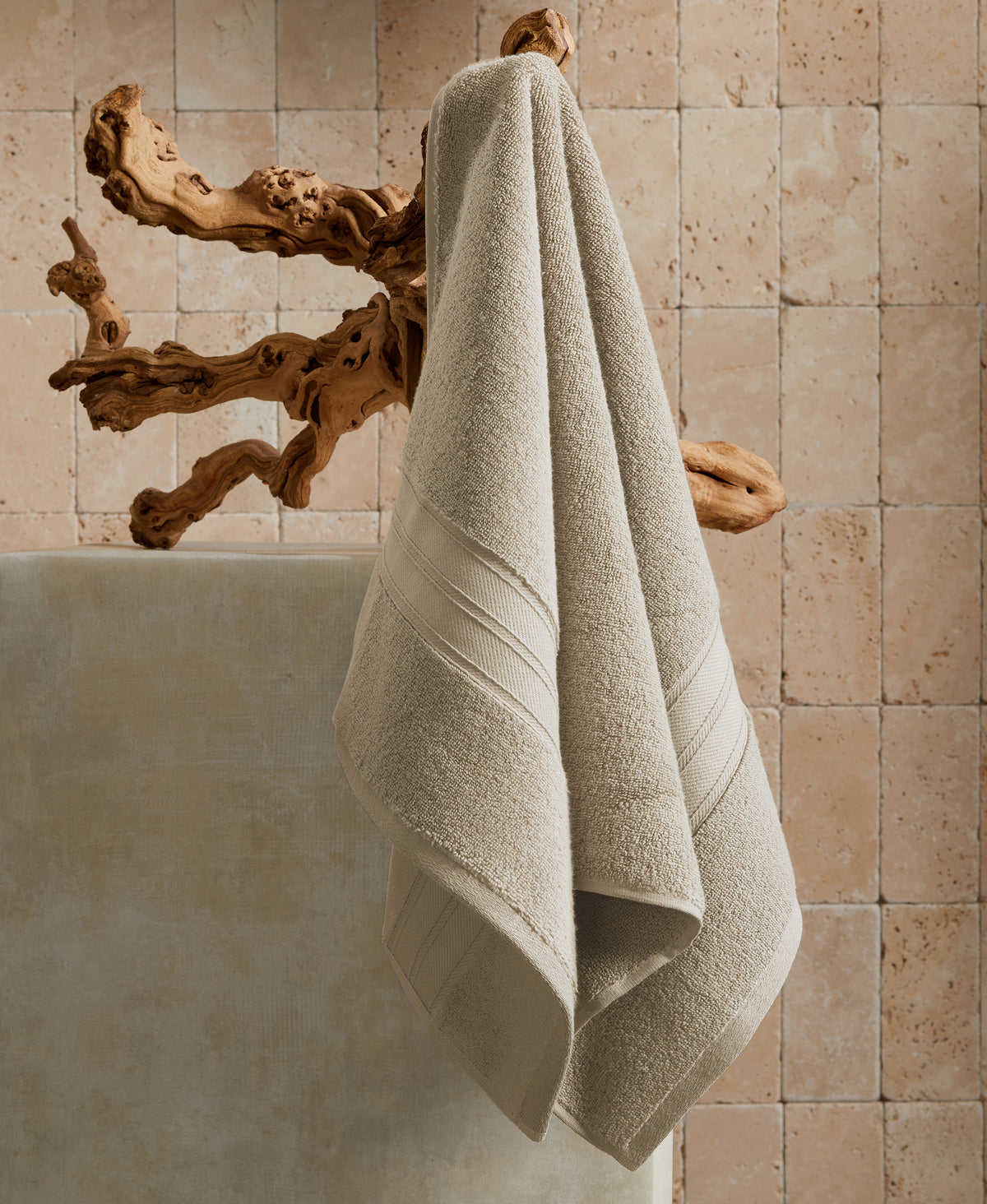 Beige towels hanging on wooden branch hook against travertine tile wall above white bathtub in spa-like bathroom