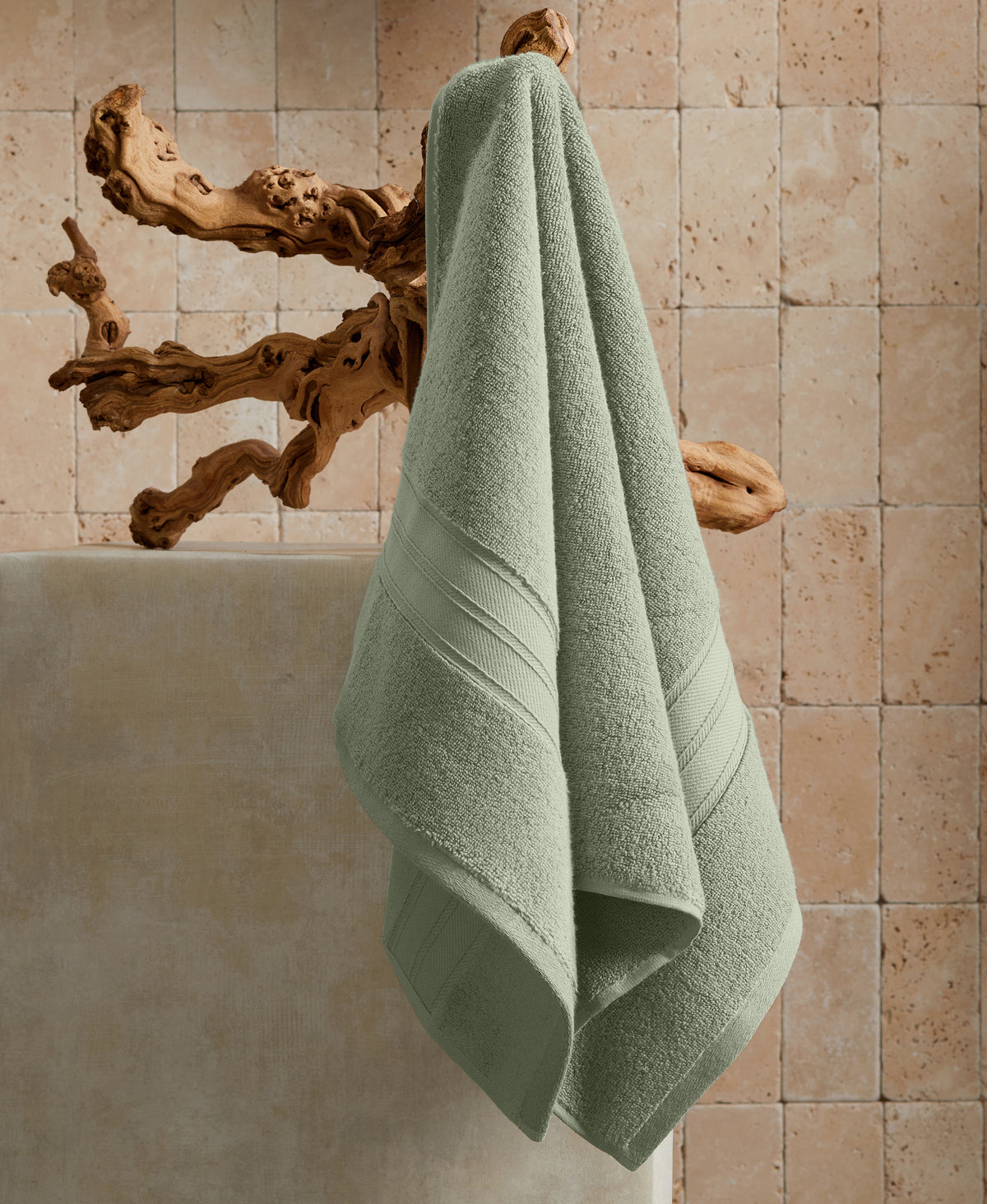 Sage green towel hanging on natural driftwood hook against beige stone tile wall above white bathtub
