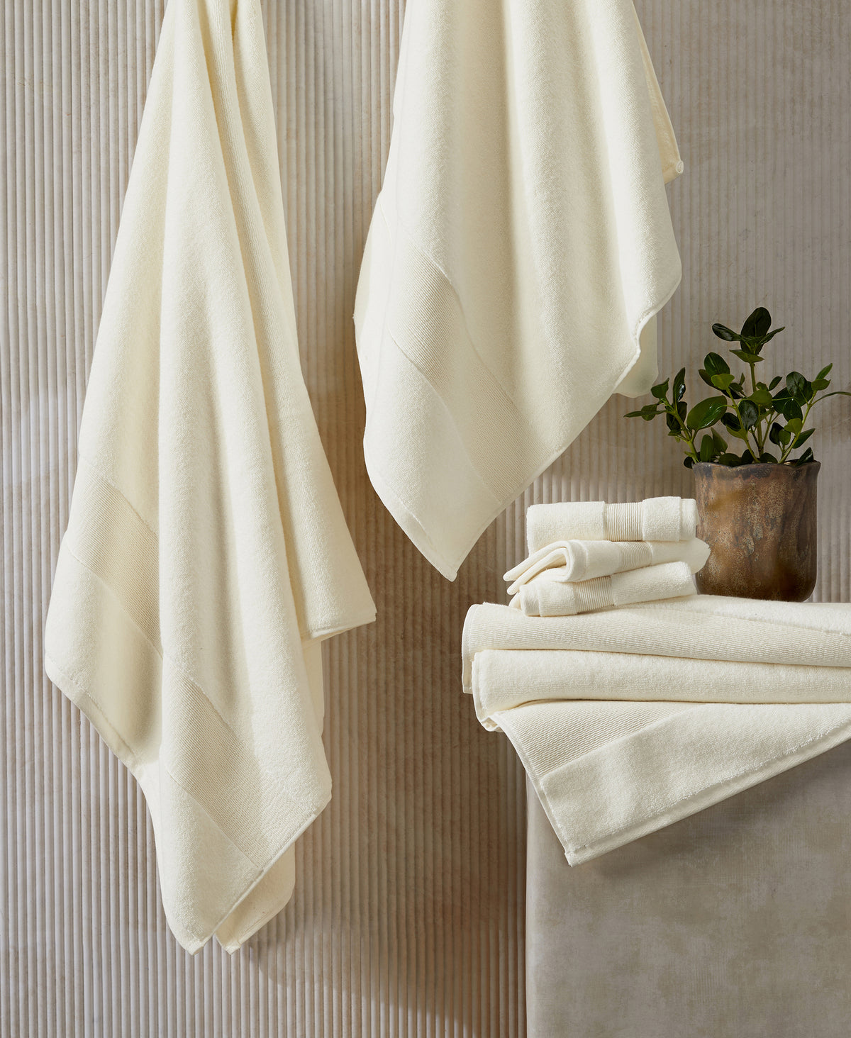 Cream-colored towels hanging on hooks and stacked on marble surface with small potted plant against textured wall