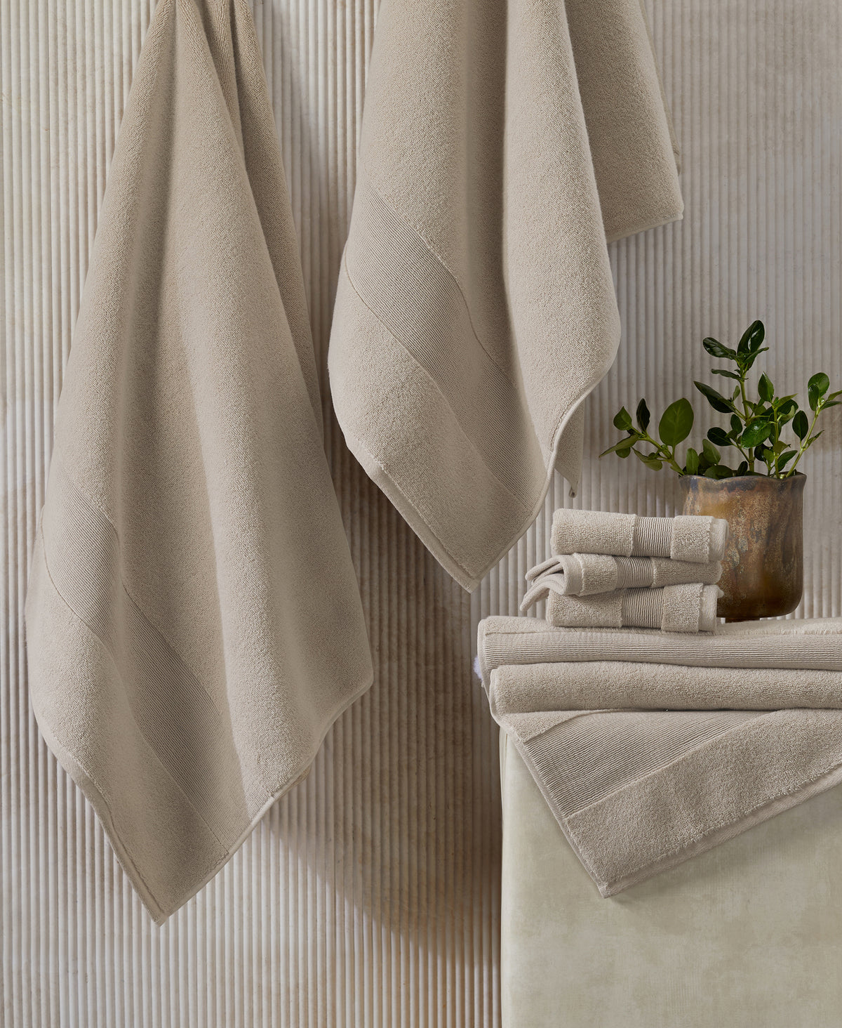 Beige towels hanging on hooks and stacked on white surface with small potted plant against textured wall