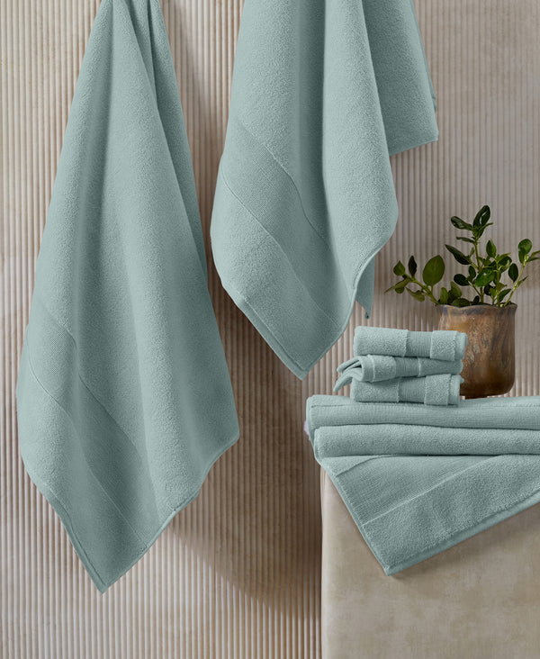 Sage green towels hanging and stacked on white surface with small potted plant against textured beige wall