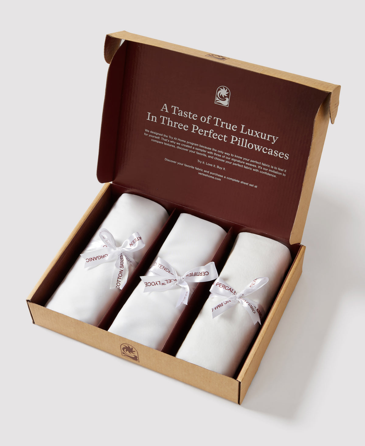 Brown and tan packaging box displaying three white rolled pillowcases with fabric name ribbons, lid open showing luxury bedding sample set.