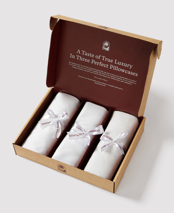 Brown and tan packaging box displaying three white rolled pillowcases with fabric name ribbons, lid open showing luxury bedding sample set.