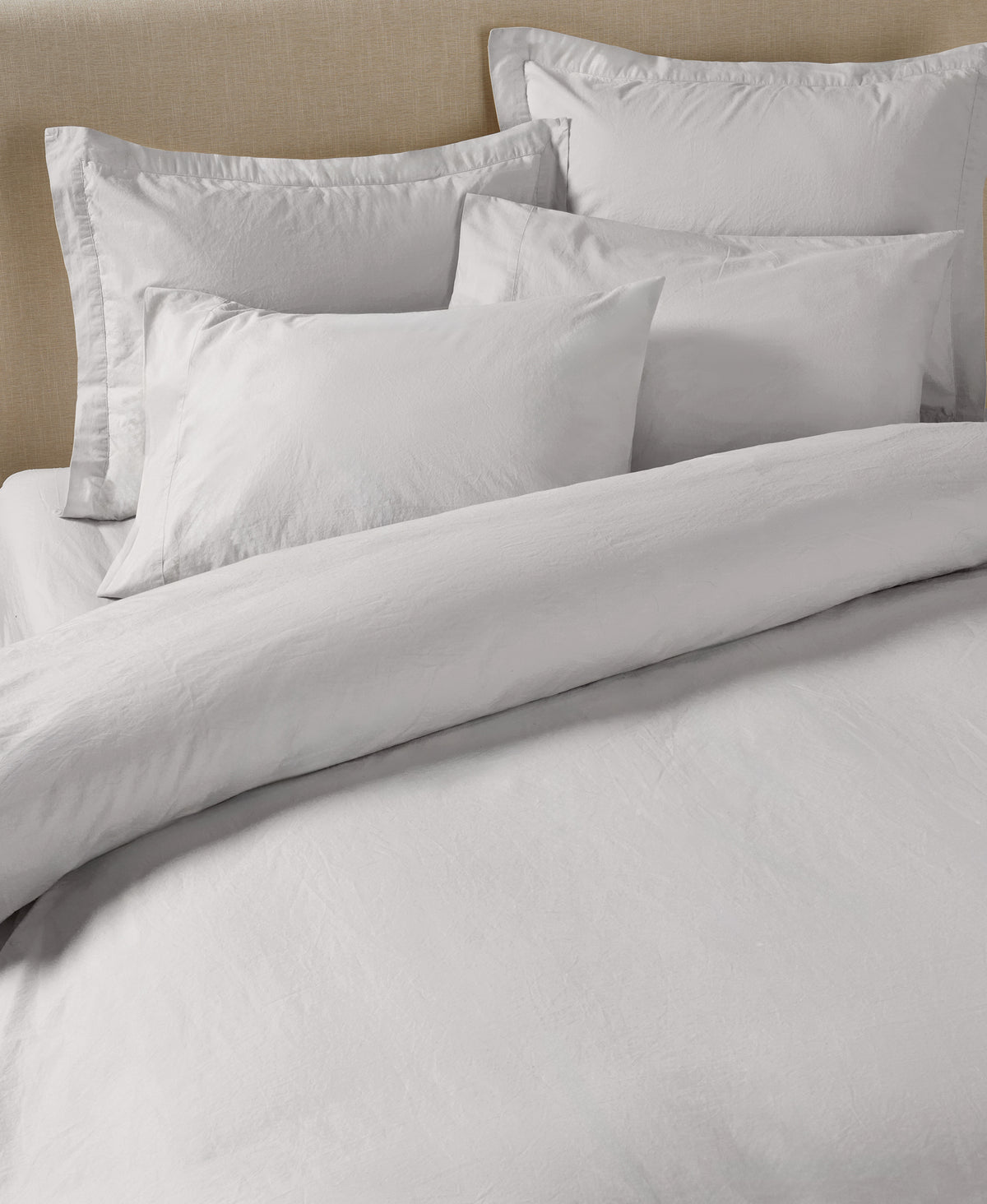 White linen bedding set with duvet cover and multiple pillows against beige upholstered headboard