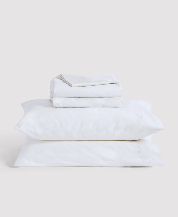 Stack of cream-colored bedding with black stripe details including folded sheets and two pillows against white background
