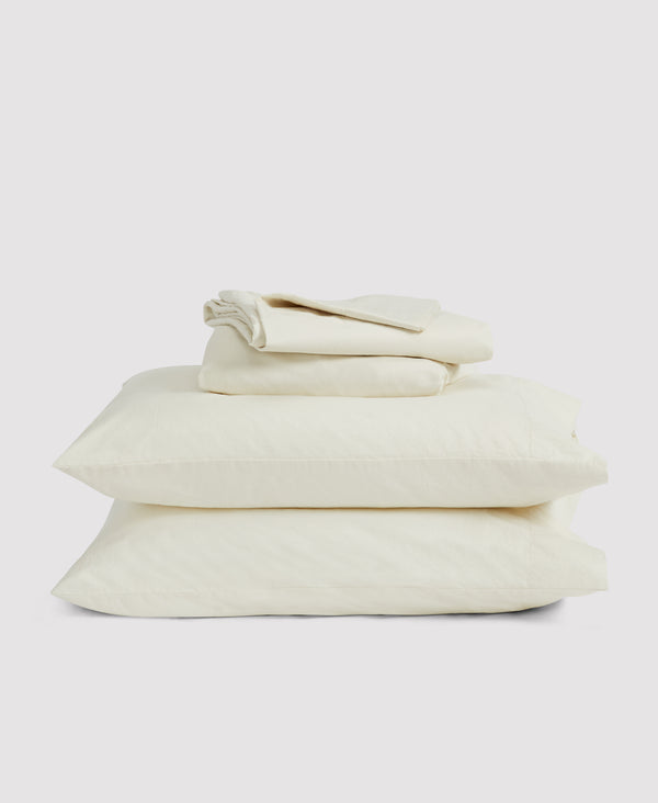 Stack of cream-colored bedding including fitted sheet, flat sheet, and two pillowcases against white background