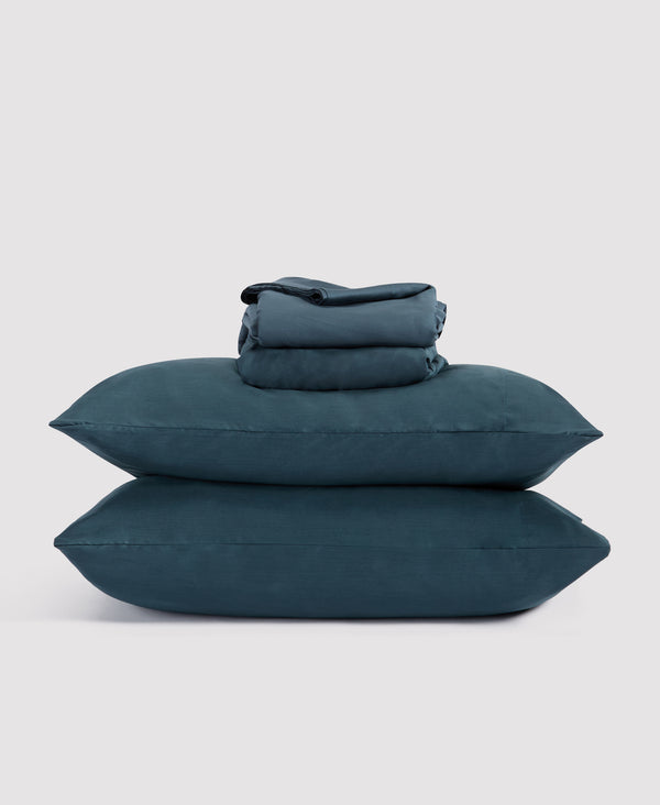 Stack of dark teal blue bedding including two pillows and folded sheets against a light gray background